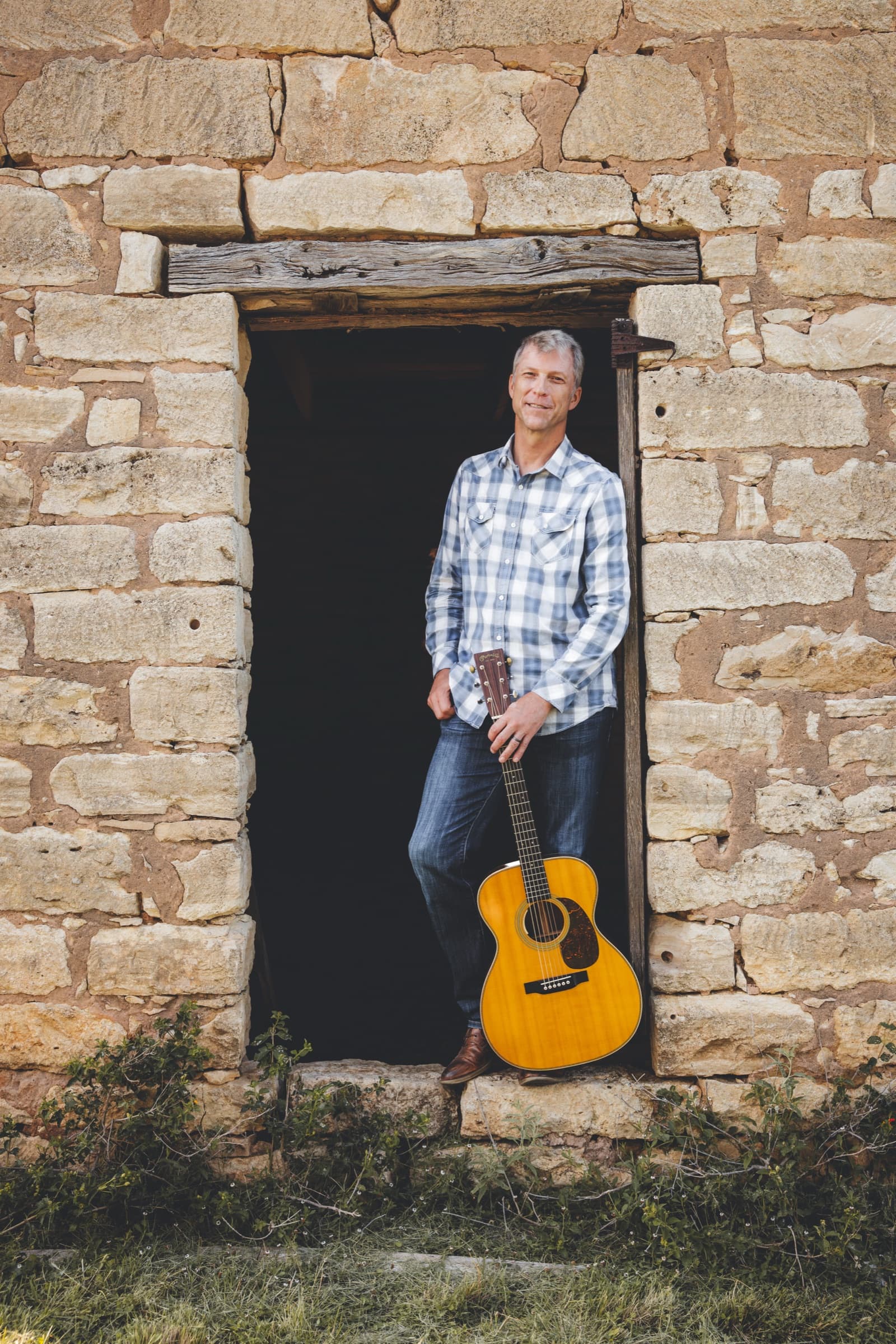 Ian Pedersen standing in a stone doorway with his guitar