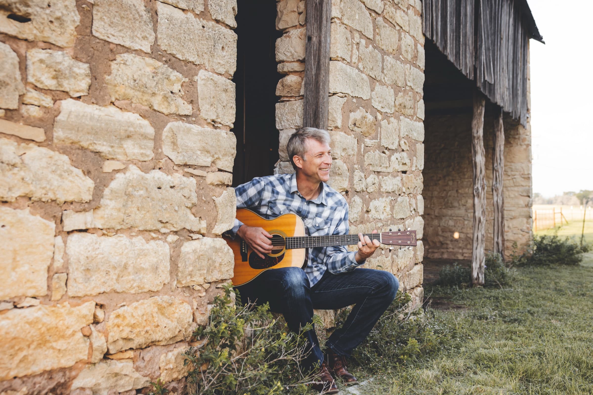 Ian Pedersen playing an acoustic guitar beside a limestone barn